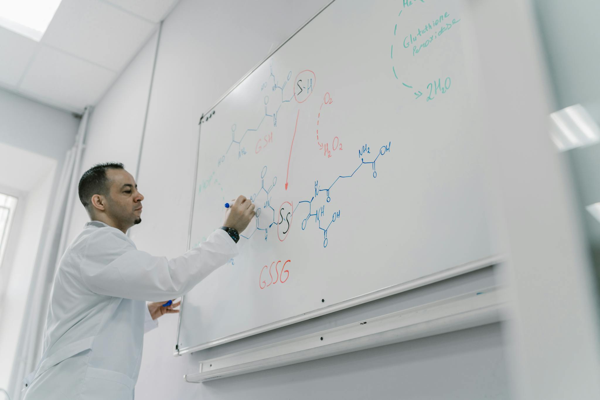 Male teacher explains chemical equation on whiteboard in science classroom setting.
